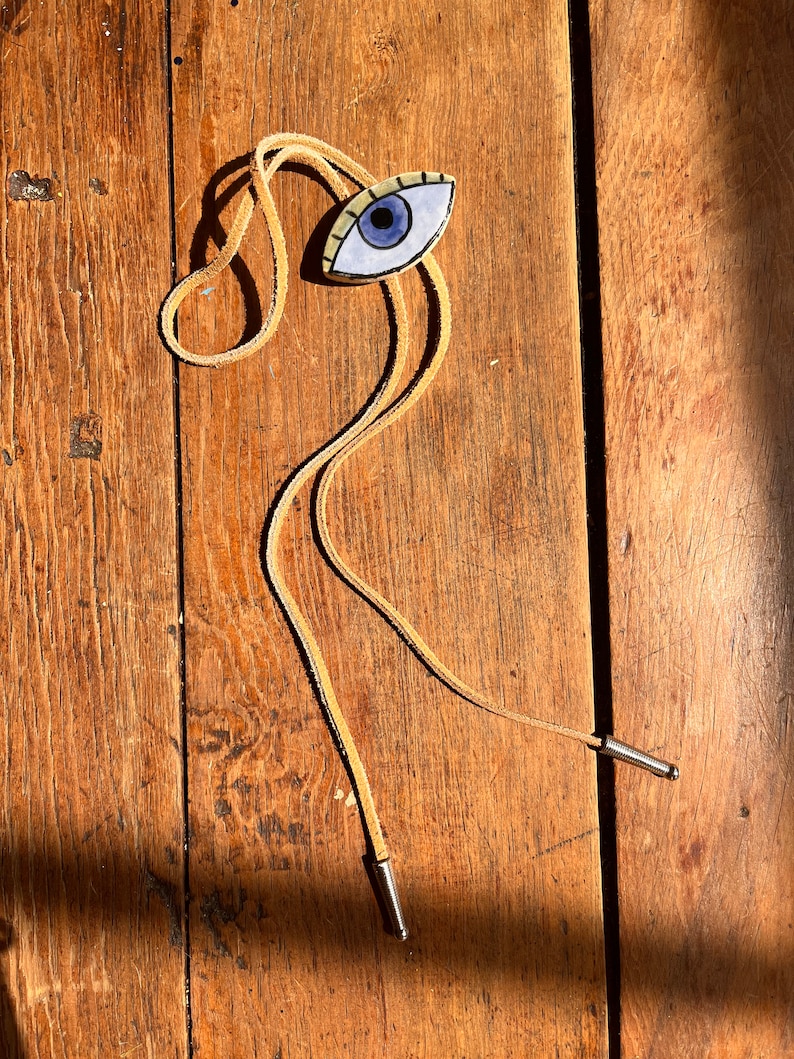 May include: A bolo tie featuring an eye-shaped pendant with a blue iris, black outline, and tan-colored lashes. The pendant is attached to a tan leather cord with silver-colored metal tips. The tie rests on a wooden surface.