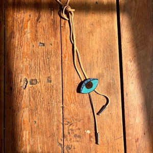 May include: A bolo tie with a light brown cord and a turquoise eye-shaped pendant. The eye has a brown iris and a black pupil. The tie has silver-colored metal tips.