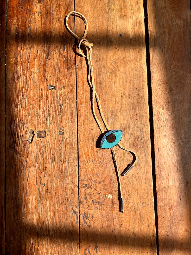 May include: A bolo tie with a light brown cord and a turquoise eye-shaped pendant. The eye has a brown iris and a black pupil. The tie has silver-colored metal tips.