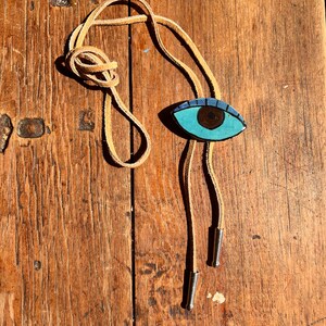 May include: A bolo tie with a hand-painted eye design. The eye is turquoise with a brown iris and a dark blue outline. The tie has a tan leather cord and silver-colored metal tips. The background is a weathered wooden surface.