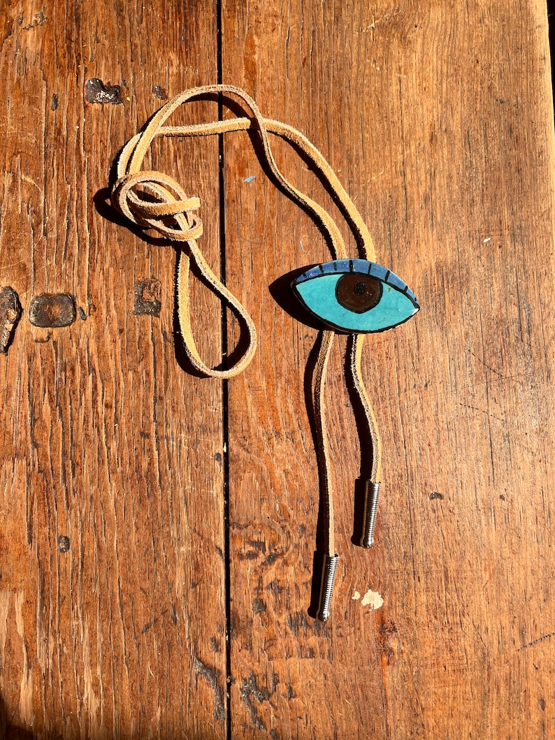 May include: A bolo tie with a hand-painted eye design. The eye is turquoise with a brown iris and a dark blue outline. The tie has a tan leather cord and silver-colored metal tips. The background is a weathered wooden surface.