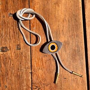 May include: A bolo tie with a gray eye-shaped pendant featuring an orange and black iris. The tie has a light tan leather cord with white accents and gold-colored metal tips. The tie is displayed on a wooden surface.