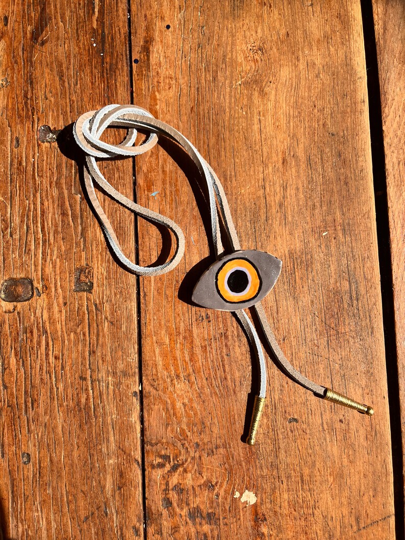 May include: A bolo tie with a gray eye-shaped pendant featuring an orange and black iris. The tie has a light tan leather cord with white accents and gold-colored metal tips. The tie is displayed on a wooden surface.