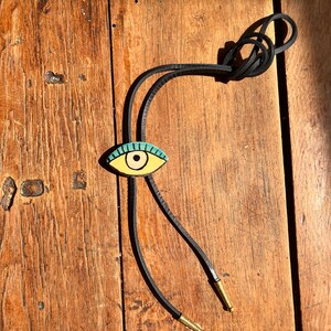 May include: A bolo tie with a gold-colored eye pendant featuring a turquoise iris and black outline. The pendant is attached to a black leather cord with gold-colored metal tips. The tie rests on a weathered wooden surface.