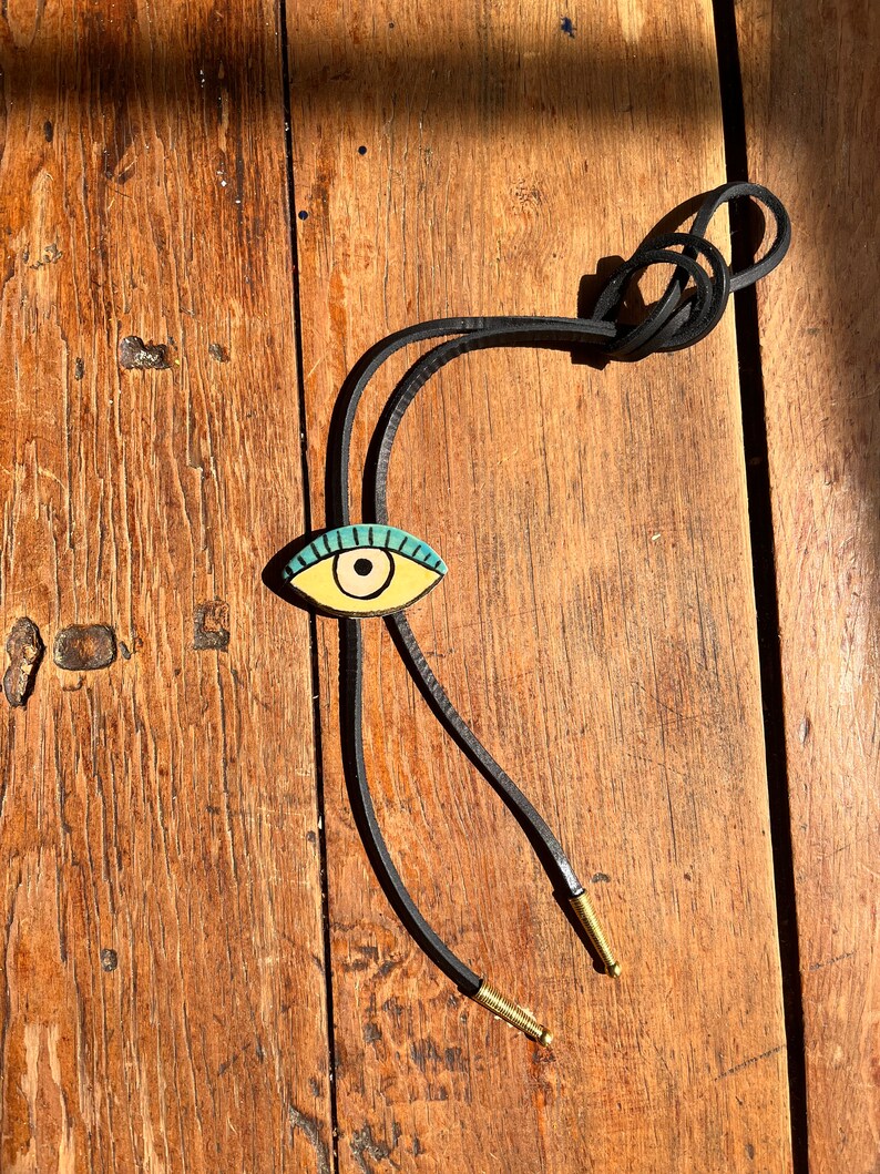 May include: A bolo tie with a gold-colored eye pendant featuring a turquoise iris and black outline. The pendant is attached to a black leather cord with gold-colored metal tips. The tie rests on a weathered wooden surface.