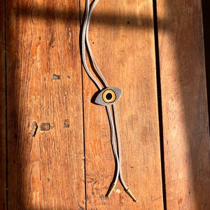 May include: A bolo tie with a gray eye-shaped pendant featuring a yellow and black iris. The cord is white with gold-colored metal tips. The tie is displayed on a wooden surface with natural light.