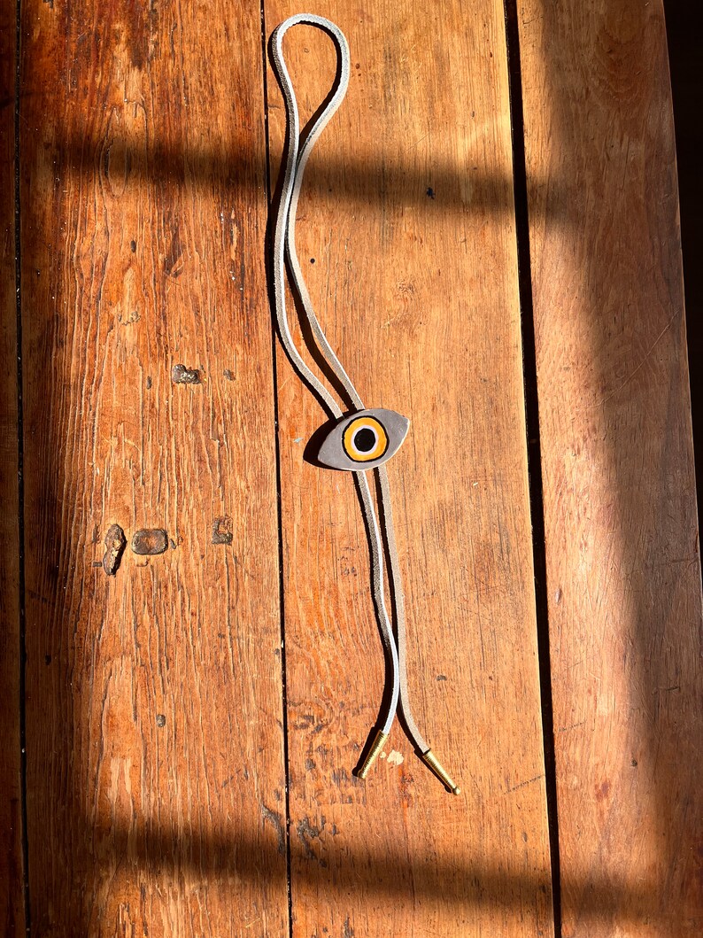 May include: A bolo tie with a gray eye-shaped pendant featuring a yellow and black iris. The cord is white with gold-colored metal tips. The tie is displayed on a wooden surface with natural light.