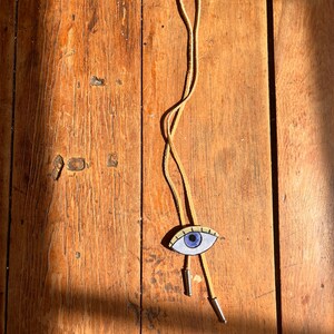 May include: A tan leather bolo tie with a painted eye pendant. The eye has a blue iris, black eyelashes, and a cream-colored background. The tie has silver-colored metal tips and is displayed on a wooden surface.