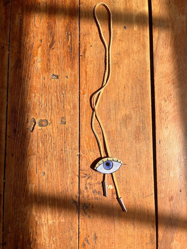 May include: A tan leather bolo tie with a painted eye pendant. The eye has a blue iris, black eyelashes, and a cream-colored background. The tie has silver-colored metal tips and is displayed on a wooden surface.