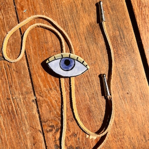 May include: A bolo tie featuring a hand-painted eye pendant with a blue iris and a tan leather cord. The eye is outlined in black and has a light yellow background. The cord has silver-colored metal tips.