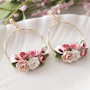 May include: A pair of gold hoop earrings adorned with a cluster of handcrafted flowers. The floral arrangement features roses and other blooms in shades of pink and white, accented with green leaves. The earrings are displayed on a white surface.