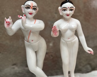 May include: Two white porcelain statues of figures with black hair and red painted details. The figures are standing on white bases.