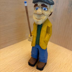 May include: A hand-carved wooden figurine of an elderly man wearing a yellow jacket, blue jeans, and a gray cap. He holds a wooden stick with a blue tip. The figure has a smiling expression and is standing on a wooden surface.