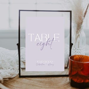 May include: A black framed sign with a light purple background. The sign says "TABLE eight" in white with a purple cursive "eight". The date "10.12.2022" and the names "Sebastian + Avery" are printed in purple below the table number.