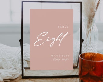 May include: A black framed table number sign with a blush pink background. The number "Eight" is written in white cursive script. The date "07.05.2022" and the names "Riley & Joseph" are written in white below the number.
