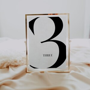 May include: A gold-framed table number with a black number three and the word "THREE" printed below it.