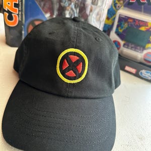 Puede incluir: Una gorra de béisbol negra con un logotipo de X-Men bordado en la parte delantera. El logotipo consiste en un borde circular amarillo que rodea un símbolo X rojo dividido en cuatro secciones triangulares. La gorra tiene una visera curva.