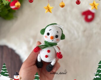 May include: A crocheted snowman toy with a green hat, white body, red mittens, and black boots. The snowman is holding a red mitten in each hand. The snowman is sitting on a white surface with a green Christmas tree and a snowman in the background.