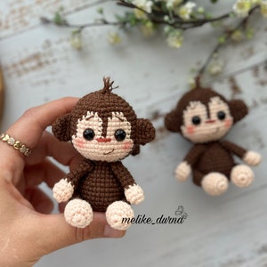 Può includere: Due giocattoli di scimmia di uncinetto marroni con occhi neri e guance rosa. Le scimmie sono sedute con le gambe incrociate.