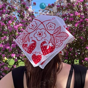 Könnte beinhalten: Weißes Bandana mit einem roten Erdbeer- und Blumenmuster. Das quadratische Bandana ist zu einem Dreieck gefaltet, wobei das Design auf den Stoff gedruckt ist. Das Bandana wird auf dem Kopf getragen.
