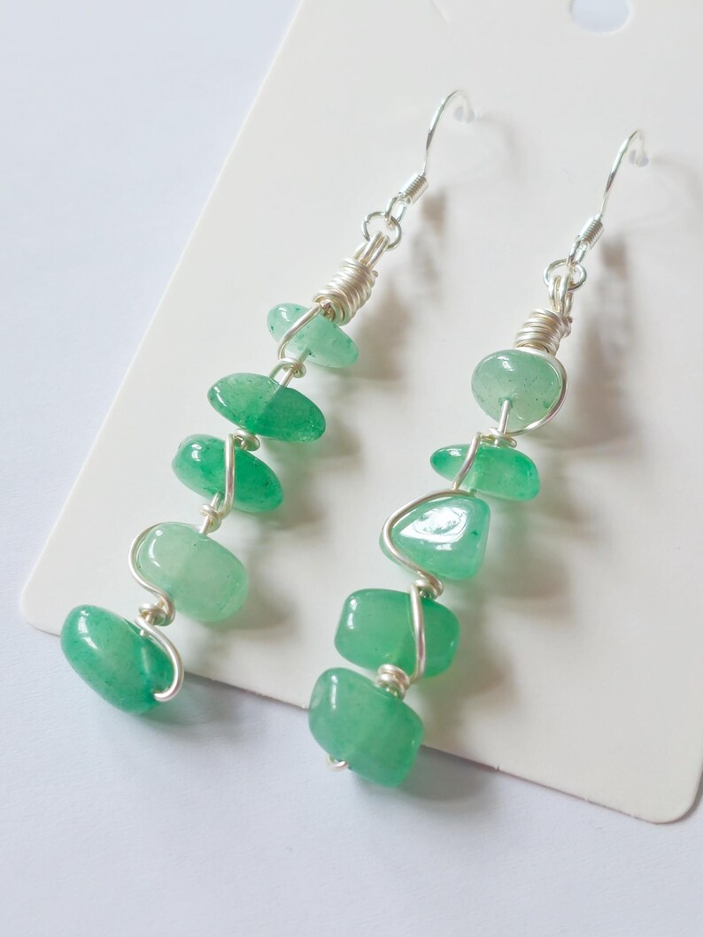 Pode incluir: Um par de brincos pendurados com contas de pedras preciosas verdes. As contas s&atilde;o enroladas em arame em um design em cascata, com um gancho prateado na parte superior. Os brincos s&atilde;o exibidos em um fundo branco.