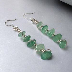 Pode incluir: Um par de brincos pendurados com contas de pedras preciosas verdes. As contas s&atilde;o enroladas em arame e suspensas de ganchos prateados. Os brincos t&ecirc;m um design simples e elegante, perfeitos para o uso di&aacute;rio.