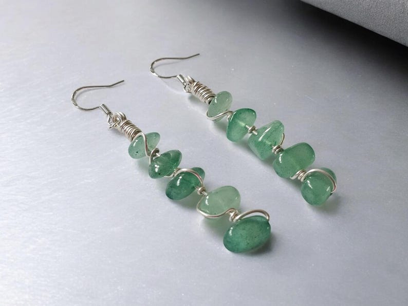 Pode incluir: Um par de brincos pendurados com contas de pedras preciosas verdes. As contas s&atilde;o enroladas em arame e suspensas de ganchos prateados. Os brincos t&ecirc;m um design simples e elegante, perfeitos para o uso di&aacute;rio.