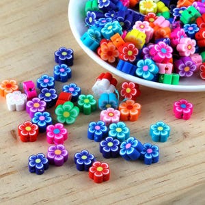 May include: Assortment of colourful flower-shaped polymer clay beads. The beads are in various colours, including red, orange, yellow, green, blue, purple, and pink. The beads are scattered on a wooden surface.