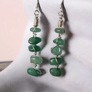 Pode incluir: Brincos pendurados com contas de pedras preciosas verdes. Cada brinco tem cinco contas ovais verdes enfiadas em arame prateado. O arame &eacute; enrolado no topo e envolve cada conta, criando um visual artesanal.