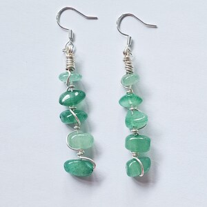 Pode incluir: Um par de brincos pendurados com pedras de aventurina verde. Cada brinco tem um gancho prateado e &eacute; composto por uma s&eacute;rie de pedras verdes polidas em forma oval, unidas por fio prateado. O fio tamb&eacute;m &eacute; enrolado em volta da parte superior das pedras.