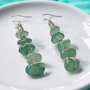 Pode incluir: Um par de brincos pendurados com contas de pedras preciosas verdes. Cada brinco tem uma s&eacute;rie de pedras verdes polidas, em forma oval, amarradas com arame prateado. Os brincos s&atilde;o exibidos em um prato branco.