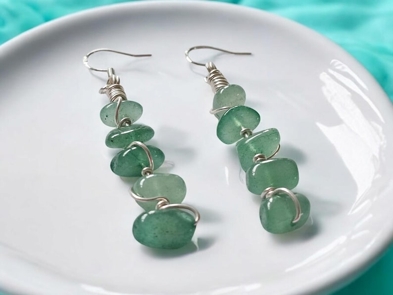 Pode incluir: Um par de brincos pendurados com contas de pedras preciosas verdes. Cada brinco tem uma s&eacute;rie de pedras verdes polidas, em forma oval, amarradas com arame prateado. Os brincos s&atilde;o exibidos em um prato branco.