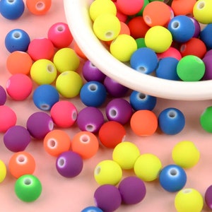 May include: A bowl filled with colorful round beads in various shades of blue, green, yellow, orange, red, and purple. The beads are scattered around the bowl on a pink surface.