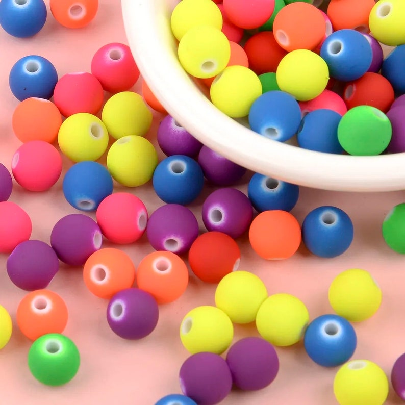 May include: A bowl filled with colorful round beads in various shades of blue, green, yellow, orange, red, and purple. The beads are scattered around the bowl on a pink surface.