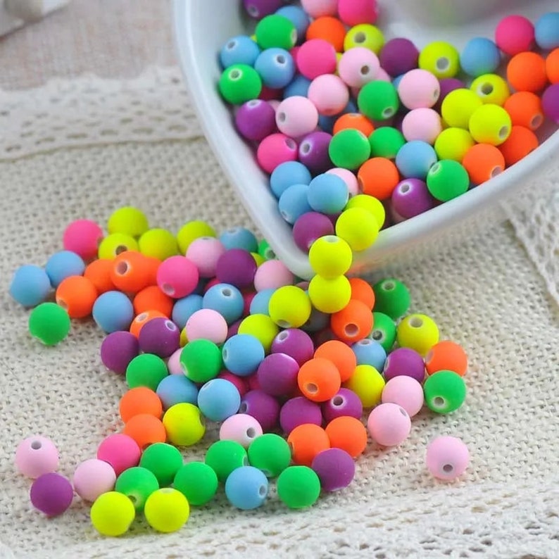 May include: A bowl filled with colorful round beads in various shades of pink, purple, blue, green, orange, and yellow. The beads are spilling out of the bowl onto a white crocheted surface.