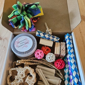 May include: A cardboard box filled with a variety of small pet toys, including a colorful knotted fleece toy, a woven straw toy, a wooden chew toy, a small jar of dandelion root treats, a natural loofah, and a few colorful woven balls.