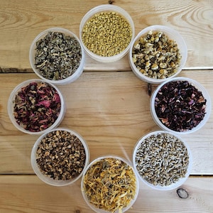 May include: Eight clear glass jars filled with different dried herbs and flowers. The jars are arranged in a circle on a wooden surface. The herbs and flowers include chamomile, lavender, rose petals, hibiscus, and calendula.