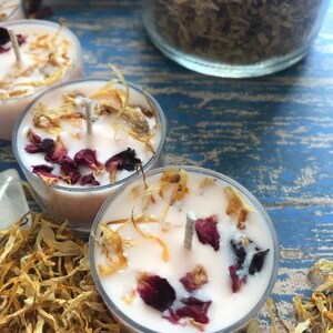 May include: Three small, round, white tea light candles with dried flower petals and a wick in the center. The candles are surrounded by dried flower petals.