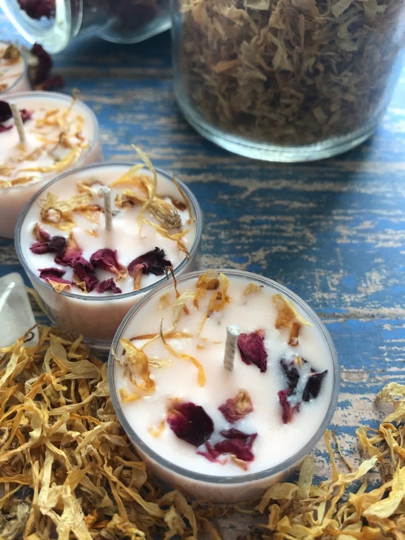 May include: Three small, round, white tea light candles with dried flower petals and a wick in the center. The candles are surrounded by dried flower petals.