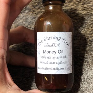 May include: A brown glass bottle with a black cap, labeled "The Burning Tree Ritual Oil Money Oil". The label also states "Made with dry herbs and botanicals under a full moon" and includes the website "BurningTreeCandle.etsy.com".