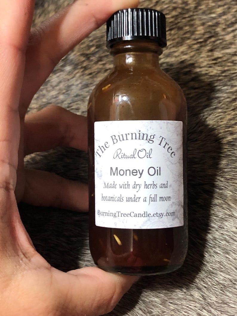 May include: A brown glass bottle with a black cap, labeled "The Burning Tree Ritual Oil Money Oil". The label also states "Made with dry herbs and botanicals under a full moon" and includes the website "BurningTreeCandle.etsy.com".