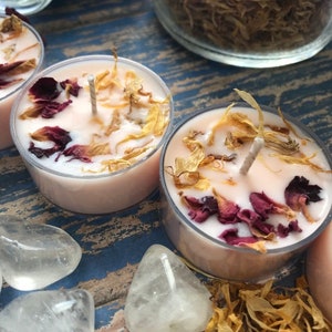 May include: Three pink tea light candles with dried flower petals and a wick in each. The candles are on a blue and brown wooden surface with white crystal hearts.