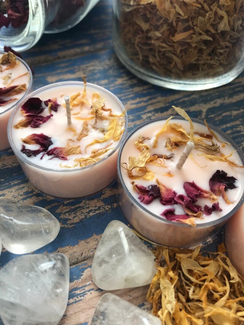 May include: Three pink tea light candles with dried flower petals and a wick in each. The candles are on a blue and brown wooden surface with white crystal hearts.