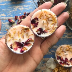 May include: Two small, round, pink tea light candles with dried rose petals and yellow flower petals embedded in the wax.