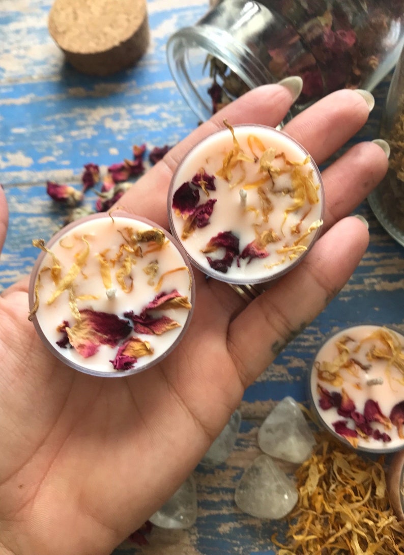 May include: Two small, round, pink tea light candles with dried rose petals and yellow flower petals embedded in the wax.