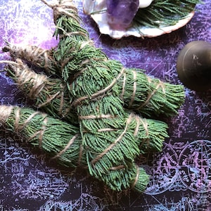 May include: Three bundles of green cedar smudge sticks tied with twine. The smudge sticks are on a dark purple background with a white and blue map pattern.