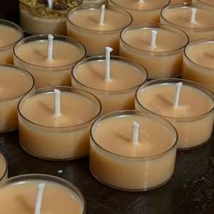Puede incluir: Primer plano de un grupo de peque&ntilde;as velas de t&eacute; redondas de color beige con mechas blancas. Las velas est&aacute;n dispuestas en filas sobre una superficie oscura.