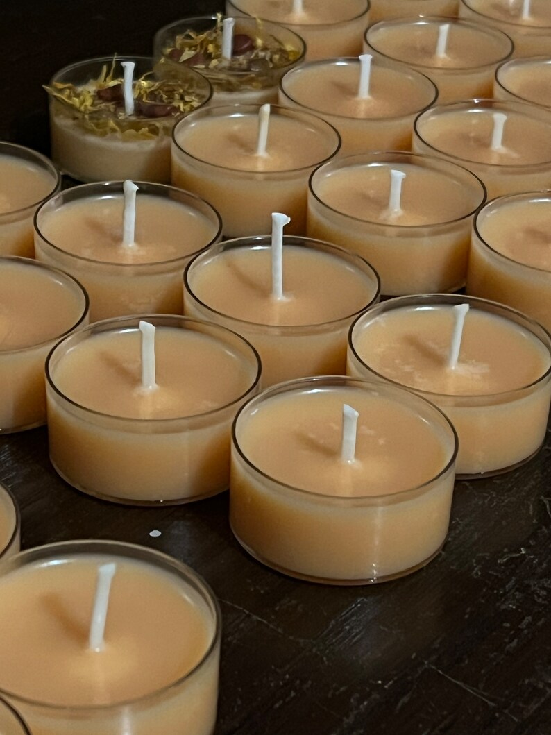 Puede incluir: Primer plano de un grupo de peque&ntilde;as velas de t&eacute; redondas de color beige con mechas blancas. Las velas est&aacute;n dispuestas en filas sobre una superficie oscura.