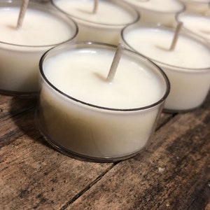 May include: A close-up of several white tea light candles in clear plastic containers. The candles are arranged in a row on a wooden surface.