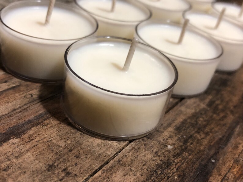 May include: A close-up of several white tea light candles in clear plastic containers. The candles are arranged in a row on a wooden surface.